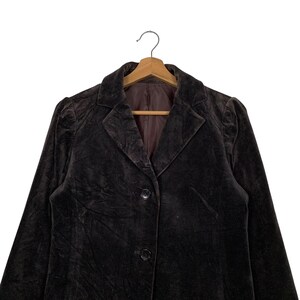 Peut inclure: Un blazer en velours marron fonc&eacute; avec un col classique et une fermeture &agrave; deux boutons. Le blazer est suspendu &agrave; un cintre en bois, mettant en valeur son design sur mesure et sa texture douce. La doublure int&eacute;rieure est de couleur marron contrastante.
