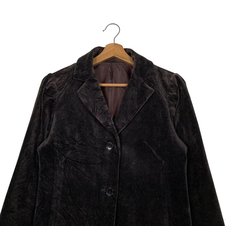 Peut inclure: Un blazer en velours marron fonc&eacute; avec un col classique et une fermeture &agrave; deux boutons. Le blazer est suspendu &agrave; un cintre en bois, mettant en valeur son design sur mesure et sa texture douce. La doublure int&eacute;rieure est de couleur marron contrastante.