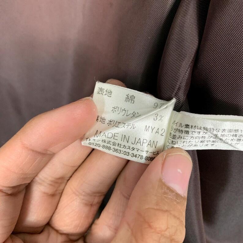 Peut inclure: Gros plan sur une &eacute;tiquette blanche avec du texte japonais et les mots "MADE IN JAPAN". L'&eacute;tiquette est tenue par une personne. L'&eacute;tiquette indique les mat&eacute;riaux : coton et polyur&eacute;thane.