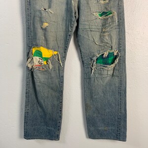 Op de afbeelding: Gedragen blauwe denim jeans met lappen stof die over de knie&euml;n zijn genaaid. De lappen zijn een groen-gele ruit en een groen-geel-rode stof met een cartoon gezicht.