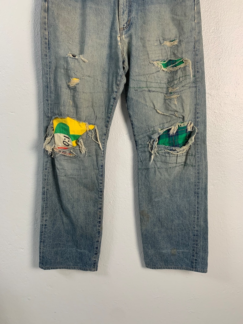 Op de afbeelding: Gedragen blauwe denim jeans met lappen stof die over de knie&euml;n zijn genaaid. De lappen zijn een groen-gele ruit en een groen-geel-rode stof met een cartoon gezicht.