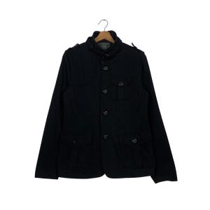 Puede incluir: Chaqueta negra de lana con cuello alzado y cierre de botones. La chaqueta tiene dos bolsillos en el pecho y dos bolsillos inferiores, y charreteras en los hombros. La chaqueta está colgada de una percha de madera.