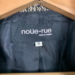 Puede incluir: Primer plano de una etiqueta negra dentro de una chaqueta. La etiqueta dice "nouie-rue" encima de "FAIRE DE CHEMIN" en texto blanco. Debajo de la etiqueta hay una etiqueta blanca con el n&uacute;mero "2". El forro de la chaqueta es negro.