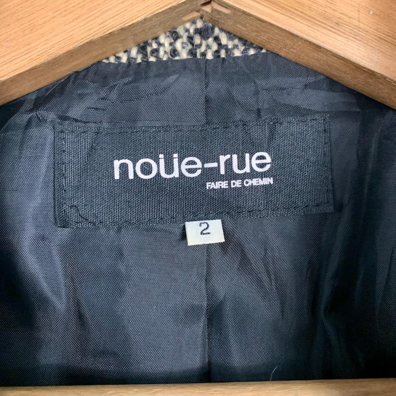 Puede incluir: Primer plano de una etiqueta negra dentro de una chaqueta. La etiqueta dice "nouie-rue" encima de "FAIRE DE CHEMIN" en texto blanco. Debajo de la etiqueta hay una etiqueta blanca con el n&uacute;mero "2". El forro de la chaqueta es negro.