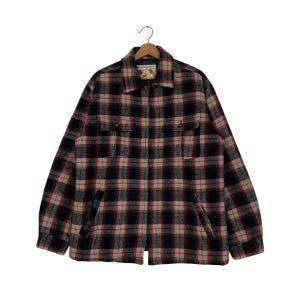 May include: A plaid jacket in a black, red, and tan color scheme, hanging on a wooden hanger. The jacket has a full zipper, two chest pockets, and two side pockets. The jacket is made of a thick, warm material.