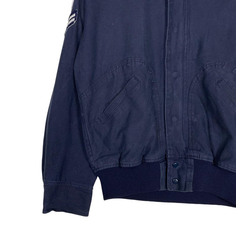 May include: Navy blue bomber jacket with a ribbed collar and cuffs. The jacket features two front pockets and a button-up closure. A white and blue emblem is visible on the upper left sleeve. The jacket is made of a durable fabric.