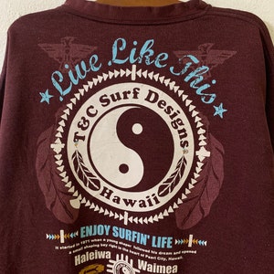 Puede incluir: Una camiseta de manga larga marr&oacute;n con un estampado gr&aacute;fico de un s&iacute;mbolo yin-yang con el texto "Live Like This" y "T&C Surf Designs Hawaii" alrededor. El texto "Enjoy Surfin' Life" est&aacute; debajo del s&iacute;mbolo con el texto "It started in 1971 when a young shaper followed his dream and opened a small shaping bay right in the heart of Pearl City, Hawaii. Haleiwa Waimea 1971" debajo.