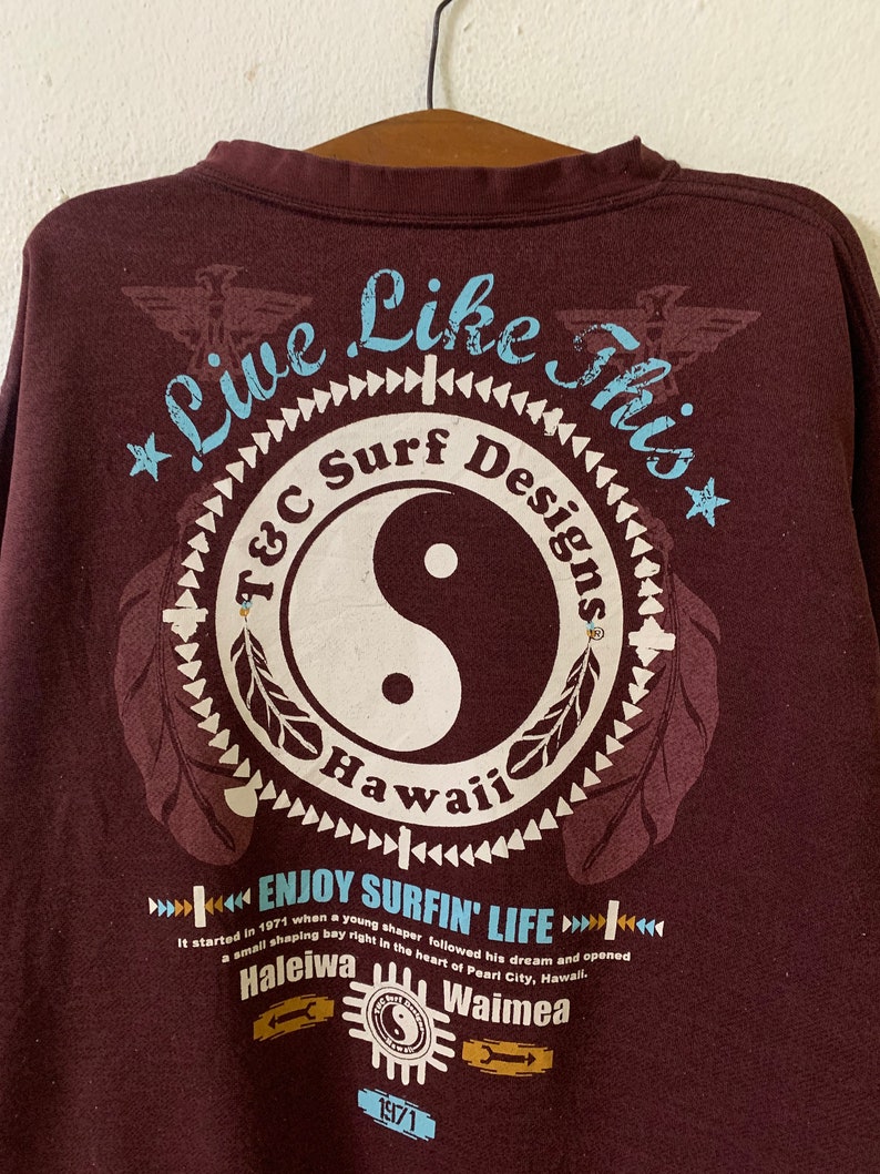 Puede incluir: Una camiseta de manga larga marr&oacute;n con un estampado gr&aacute;fico de un s&iacute;mbolo yin-yang con el texto "Live Like This" y "T&C Surf Designs Hawaii" alrededor. El texto "Enjoy Surfin' Life" est&aacute; debajo del s&iacute;mbolo con el texto "It started in 1971 when a young shaper followed his dream and opened a small shaping bay right in the heart of Pearl City, Hawaii. Haleiwa Waimea 1971" debajo.