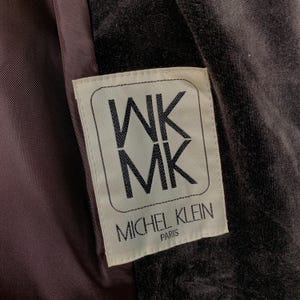 Peut inclure: Gros plan sur une &eacute;tiquette de v&ecirc;tement. L'&eacute;tiquette rectangulaire aux coins arrondis affiche les lettres "WK MK" empil&eacute;es verticalement dans un cadre. Le texte "MICHEL KLEIN PARIS" est en dessous. Le fond est un tissu sombre et textur&eacute;.