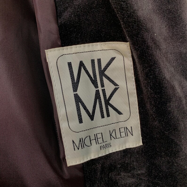 Peut inclure: Gros plan sur une &eacute;tiquette de v&ecirc;tement. L'&eacute;tiquette rectangulaire aux coins arrondis affiche les lettres "WK MK" empil&eacute;es verticalement dans un cadre. Le texte "MICHEL KLEIN PARIS" est en dessous. Le fond est un tissu sombre et textur&eacute;.