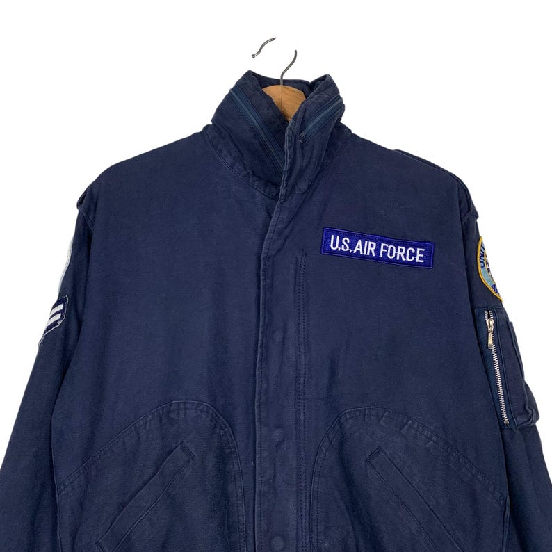 May include: Navy blue U.S. Air Force jacket with a high collar and multiple pockets. The jacket features a patch with the text "U.S. AIR FORCE" and a zipper on the sleeve. The jacket is hanging on a hanger.