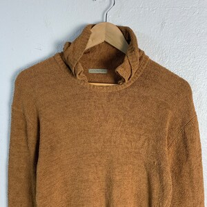 Puede incluir: Un su&eacute;ter de cuello alto marr&oacute;n con un cuello ligeramente fruncido. El su&eacute;ter est&aacute; hecho de una tela suave y texturizada.