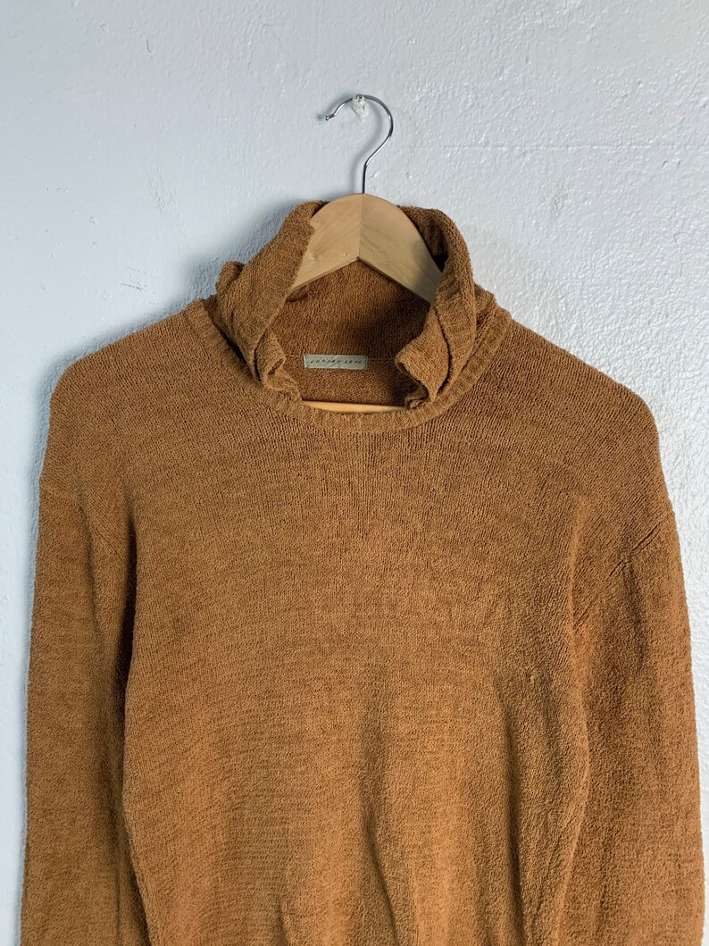 Puede incluir: Un su&eacute;ter de cuello alto marr&oacute;n con un cuello ligeramente fruncido. El su&eacute;ter est&aacute; hecho de una tela suave y texturizada.