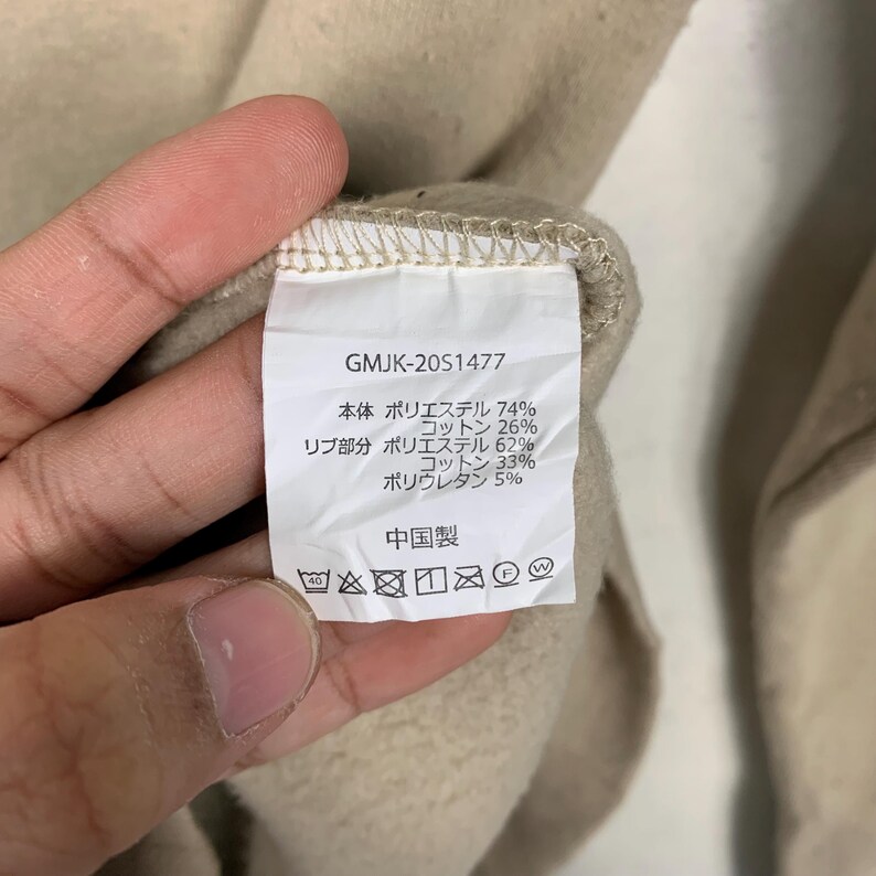 May include: A white tag with black text detailing the fabric composition of a garment. The tag reads "GMJK-2051477", "本体 ポリエステル 74% コットン 26%", "リブ部分 ポリエステル 62% コットン 33% ポリウレタン 5%", and "中国製".