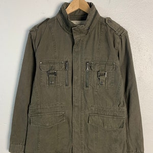 Puede incluir: Una chaqueta utilitaria verde oscuro con múltiples bolsillos y cierres de cremallera. La chaqueta tiene un cuello alto y un cierre de botones en la parte delantera.