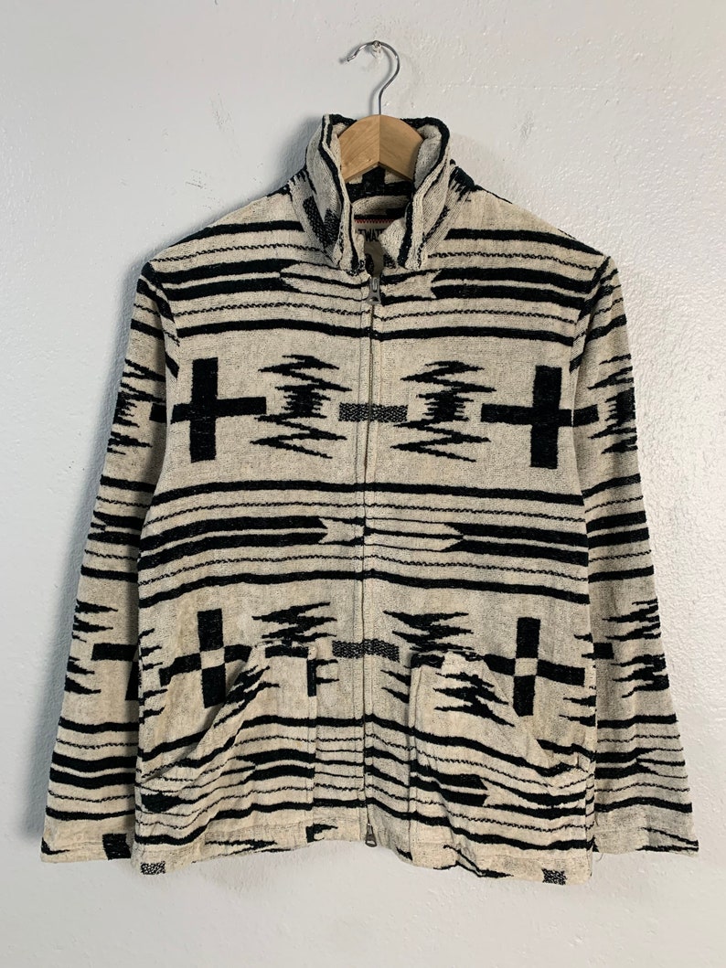 Puede incluir: Una chaqueta con cremallera y cuello alto con estampado en blanco y negro. La chaqueta tiene un dise&ntilde;o geom&eacute;trico con rayas y cruces en blanco y negro.