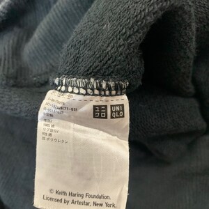 May include: A white tag with black text attached to a black sweatshirt. The tag includes the text "Keith Haring Foundation. Licensed by Artestar, New York." and a Uniqlo logo.