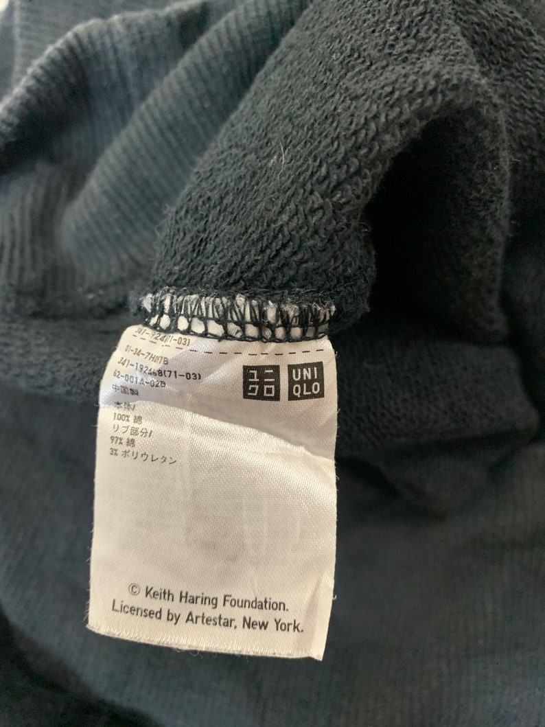 May include: A white tag with black text attached to a black sweatshirt. The tag includes the text "Keith Haring Foundation. Licensed by Artestar, New York." and a Uniqlo logo.