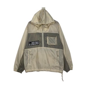 May include: A beige hooded jacket with a zipper closure and a patch that says "BESCHWA" with an alien logo. The jacket has a large pocket on the front and a drawstring at the bottom.