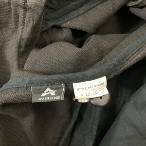 Puede incluir: Primer plano de una prenda gris oscuro con una etiqueta negra con el logotipo de Avirex. Una etiqueta blanca muestra "STYLE NO. 612209" y s&iacute;mbolos de cuidado. La tela parece ser un material de punto texturizado.