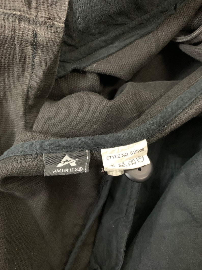 Puede incluir: Primer plano de una prenda gris oscuro con una etiqueta negra con el logotipo de Avirex. Una etiqueta blanca muestra "STYLE NO. 612209" y s&iacute;mbolos de cuidado. La tela parece ser un material de punto texturizado.