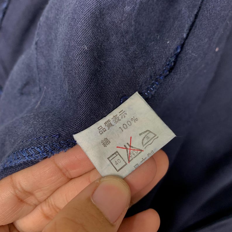 May include: Close-up of a navy blue garment with a white tag. The tag has Japanese text and symbols, including "100%" and washing instructions. The garment's fabric is visible in the background.