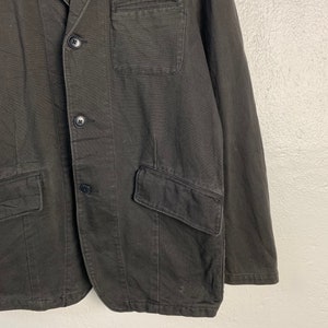 Puede incluir: Un blazer gris oscuro con cierre cl&aacute;sico de dos botones. La chaqueta presenta solapas con muescas, un bolsillo en el pecho y dos bolsillos delanteros con solapa. La tela parece ser una mezcla resistente de algod&oacute;n o lino.