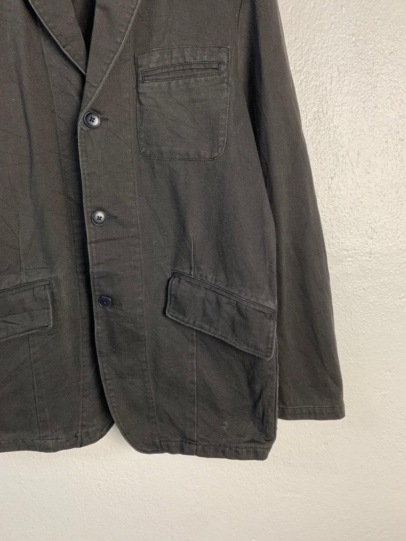 Puede incluir: Un blazer gris oscuro con cierre cl&aacute;sico de dos botones. La chaqueta presenta solapas con muescas, un bolsillo en el pecho y dos bolsillos delanteros con solapa. La tela parece ser una mezcla resistente de algod&oacute;n o lino.