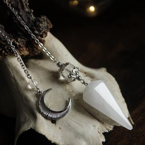 May include: A silver-toned metal chain necklace featuring a pointed, faceted white crystal pendant, a clear round bead with ornate silver-toned metal caps, and a small silver-toned crescent moon charm with textured line details along the curve.
