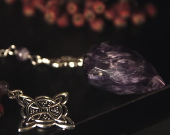 Op de afbeelding: Een gefacetteerde paarse amethist kristallen pendel bevestigd aan een zilverkleurige metalen ketting. De ketting is voorzien van een kleine ronde amethist kraal en een zilverkleurige metalen bedel met een complex, symmetrisch knoopontwerp.