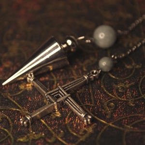 May include: A silver-toned metal pendulum with a pointed conical shape and a silver-toned metal cross charm featuring a woven-style design. The items are attached to a thin metal chain accented with two small, round, pale grey beads.