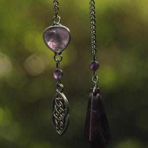 Peut inclure: Deux pendentifs suspendus à des chaînes argentées. Le pendentif de gauche présente une pierre violette claire en forme de cœur au-dessus d'une breloque en métal argenté avec un motif de nœud tressé. Le droit est un cristal violet foncé.