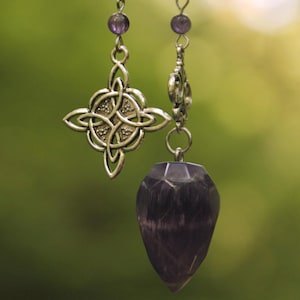 May include: Two pendulums hang against a blurred green backdrop. One has a silver Celtic knot pendant, the other a faceted, teardrop-shaped amethyst crystal. Both are suspended by delicate silver chains with purple beads.