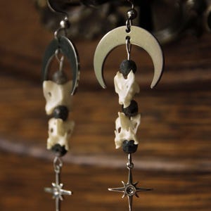 Può includere: Un paio di orecchini pendenti con un ciondolo a mezzaluna color argento in alto, seguito da un'alternanza di piccole perle porose grigio scuro e segmenti color bianco sporco, che terminano con un ciondolo a stella a quattro punte.