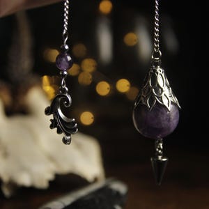 Può includere: Due ciondoli in metallo color argento appesi a catenelle. Uno presenta una pietra sferica viola sormontata da un cappuccio decorativo in metallo e una base a punta. L'altro è composto da una piccola perla viola e un ciondolo a spirale.