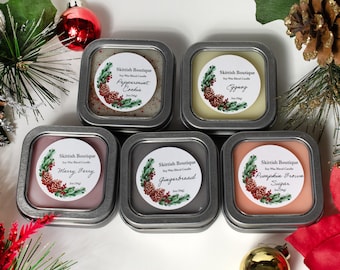 May include: Five small, square, silver tins with lids containing soy wax blend candles. Each tin has a white label with a watercolor design of a pine cone and greenery. The labels also include the text "Skittish Boutique" and the name of the scent: Peppermint Cookie, Eggnog, Merry Berry, Gingerbread, and Pumpkin Brown Sugar.