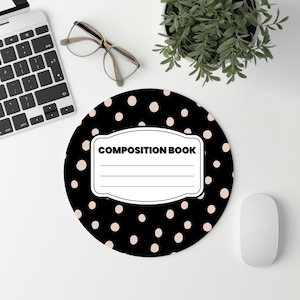 May include: A round black mousepad with a polka dot pattern and the words "COMPOSITION BOOK" in a white label. A white computer mouse, keyboard, glasses, and a small plant are also visible.