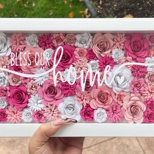 May include: A white frame with a glass front filled with pink and white paper flowers. The flowers are arranged in a dense pattern and the text "Bless our home" is written in white cursive lettering.