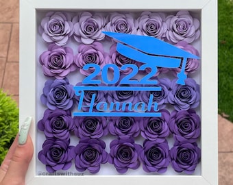 May include: A white shadow box with a graduation cap and the year 2022 in blue lettering. The name "Hannah" is also in blue lettering. The box is filled with purple paper flowers.