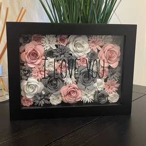 May include: A black framed shadow box with a floral arrangement of pink and gray paper flowers. The flowers are arranged in a random pattern and the words "I LOVE YOU" are printed in black on the glass.