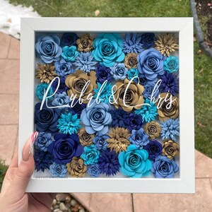May include: A white frame with a blue, gold, and teal paper flower arrangement inside. The names "Ryb&Chris" are written in white script in the center of the arrangement.
