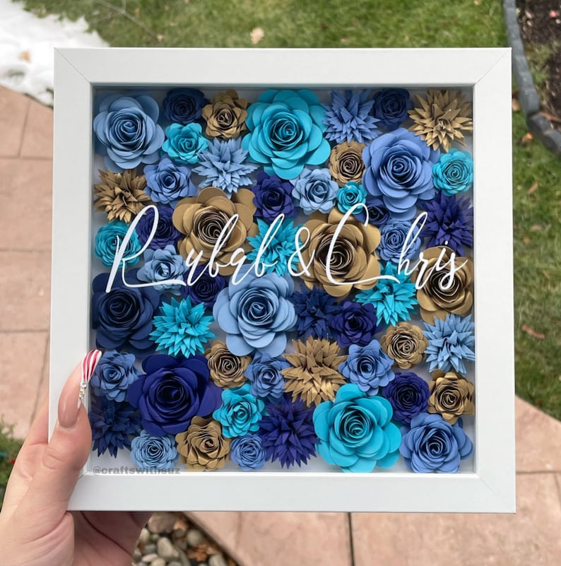 May include: A white frame with a blue, gold, and teal paper flower arrangement inside. The names "Ryb&Chris" are written in white script in the center of the arrangement.