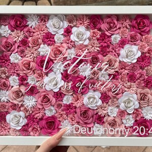 May include: A framed artwork featuring a pink and white paper flower arrangement with the text "Victoria goddess of victory - Deuteronomy 20:4" in white lettering.