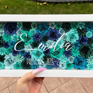 May include: A white frame filled with teal, blue, and green paper flowers. The name "Emilia" is written in white cursive script above the text "Proverbs 31:25-26".