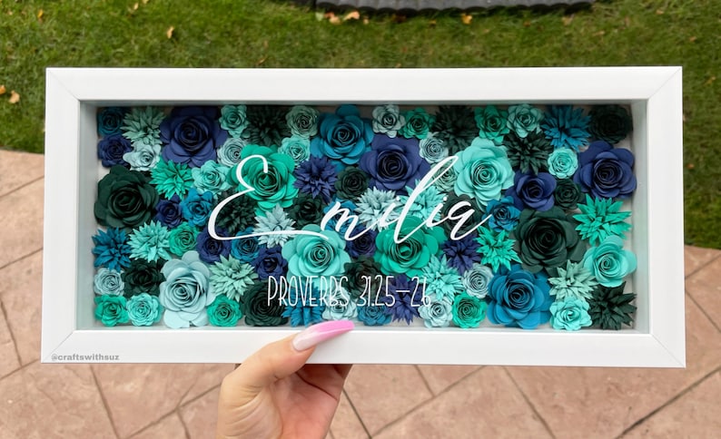 May include: A white frame filled with teal, blue, and green paper flowers. The name "Emilia" is written in white cursive script above the text "Proverbs 31:25-26".