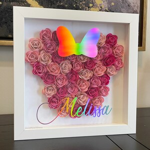 May include: A white frame with a pink and white paper flower arrangement in the shape of a mouse head. A rainbow bow is on top of the flowers. The name "Melissa" is written in a rainbow font below the flowers.