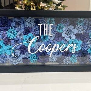 May include: A black framed shadow box with a blue and teal paper flower arrangement. The text "THE Coopers" is displayed in white lettering.