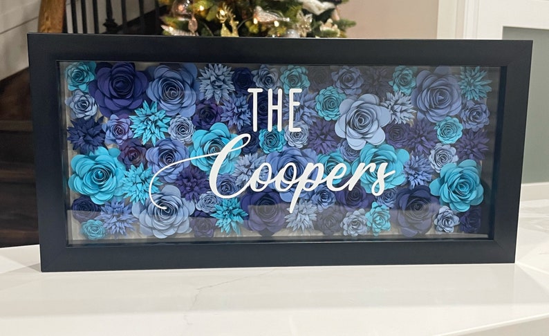 May include: A black framed shadow box with a blue and teal paper flower arrangement. The text "THE Coopers" is displayed in white lettering.