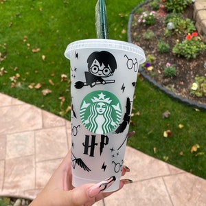 May include: A white reusable cup with a black and green Starbucks logo and Harry Potter themed graphics, including a character, a broom, and the letters HP.