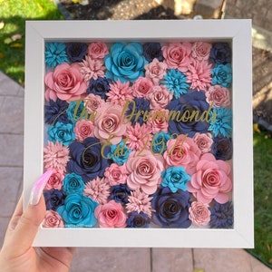 May include: A white frame with a glass front filled with paper flowers in shades of pink, blue, and navy. The flowers are arranged in a dense pattern and the text "The Drummonds Est. 2011" is written in gold lettering.